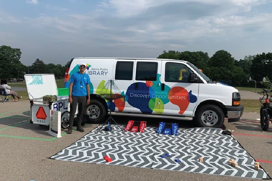 BPL staff member stands in front of the van with an outdoor carpet and activities set up
