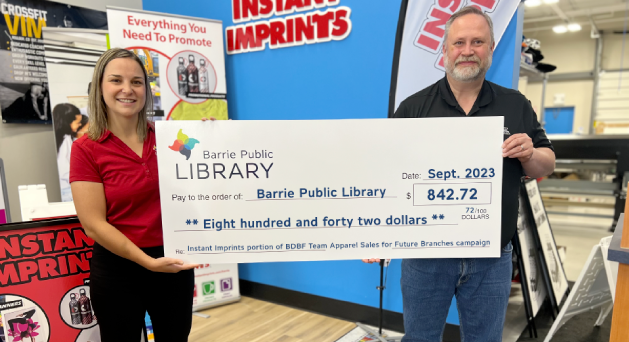 Bruce McGillvary from Instant Imprints stands with Katelyn Lees from the Library, holding a large-sized cheque for $842 dollars