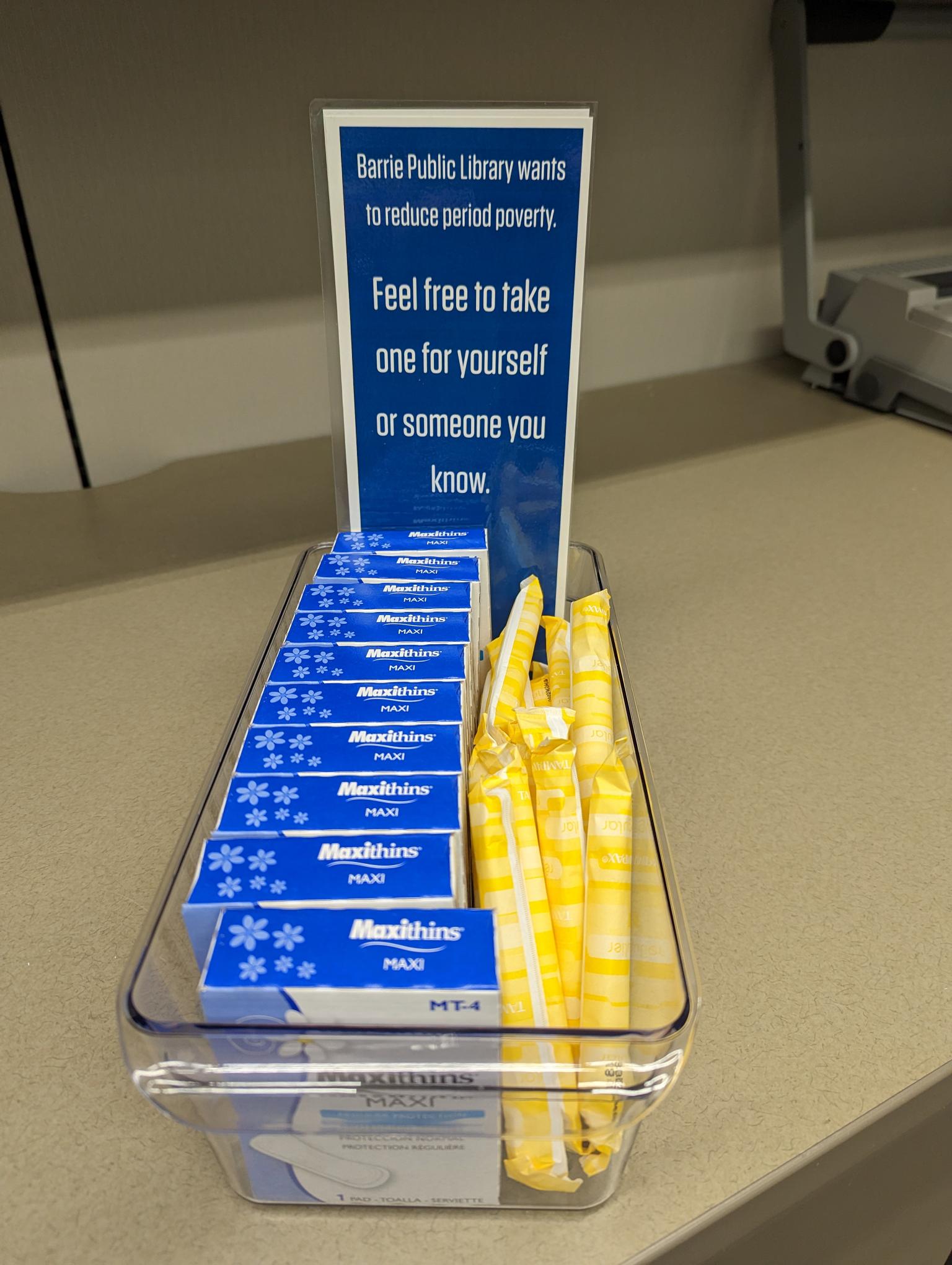 "Barrie Public Library wants to reduce period poverty. Feel free to take one for yourself or for someone you know." Sign sits over a bin with boxes of pads and tampons.